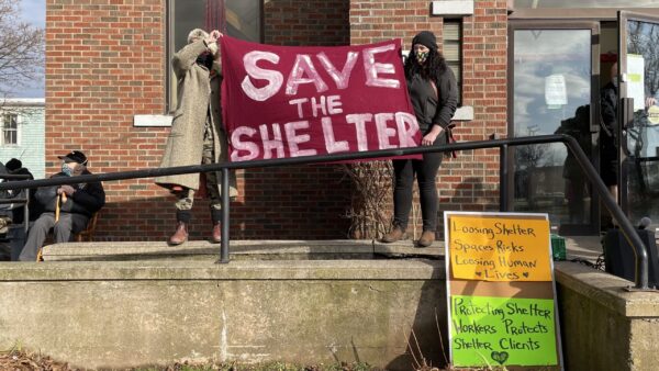 两名参加集会的人站在哈利法克斯北公园街友谊中心避难所门外的栏杆上。他们举着写着“拯救我们的庇护所”的牌子。