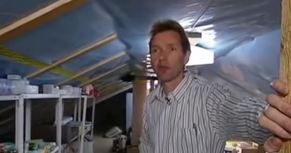 一个白人在阁楼上，可能是在房子里。天花板衬着塑料。他身后是摇摇晃晃的塑料或金属架子，上面零星地放着小包装的东西。你可以看到他身后的地板上有几箱东西。