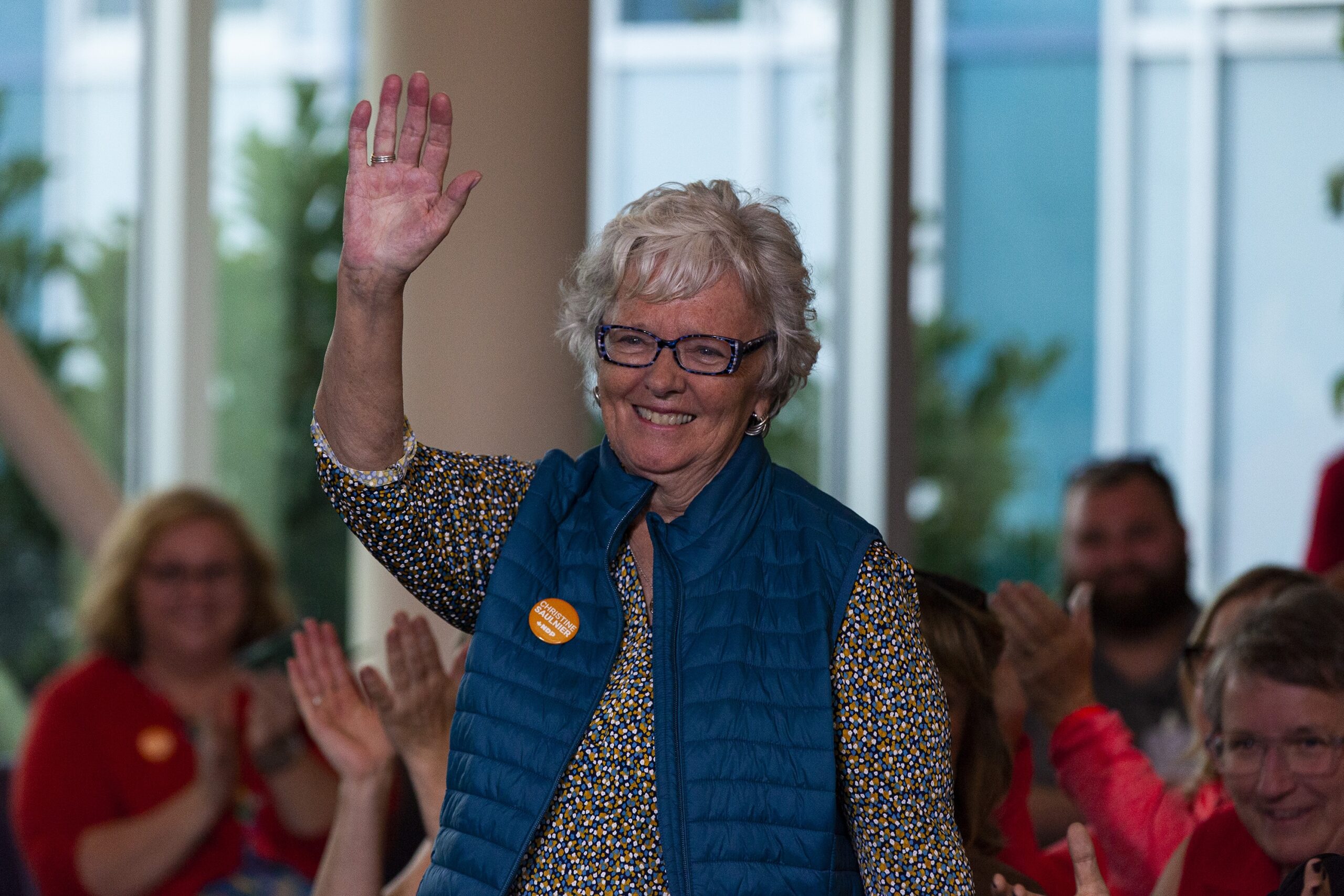 一名身穿蓝色背心、别着橙色别针的妇女向人群挥手。在背景中，人们鼓掌。