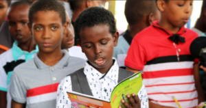 一个年轻的黑人男孩在一群其他男孩靠近相机，从一本书朗读。