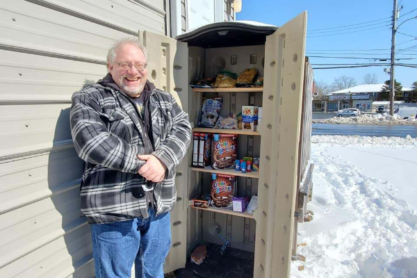 戴眼镜、灰发的白人男子大卫·阿尔德斯站在食品储藏室敞开的门口，开心地笑着。里面的架子上有几样东西，包括烤面包、盒装麦片和罐头食品。