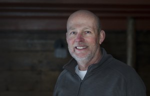 一名身穿深灰色拉链毛衣和白色t恤的男子微笑着站在一间镶着木板的房间里。