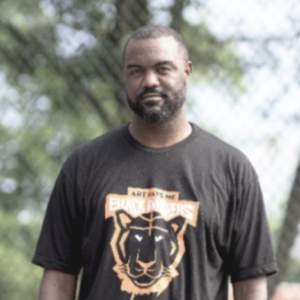 杜安·琼斯(Duane Jones)，一个穿着正面印有老虎图案的黑色t恤的黑人男子。他站在一个夏日的铁链围栏前，在他身后，你可以看到背景中模糊的绿色树木。