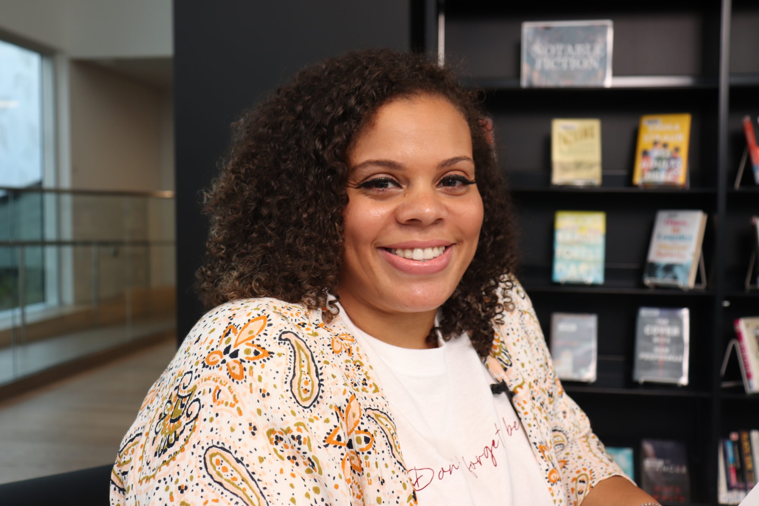 身穿浅色衣服的黑人女士对着镜头微笑，背景是书架上的书。