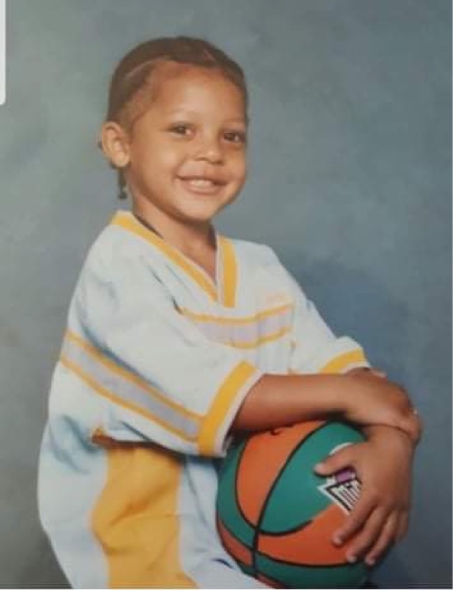 一个长着玉米脑的黑人小男孩拿着一个成人大小的绿色和橙色篮球，在一张肖像照片中微笑着