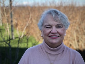 一个有着较短的灰色波浪状头发的女人，戴着珍珠耳环，穿着淡粉色的高领毛衣。