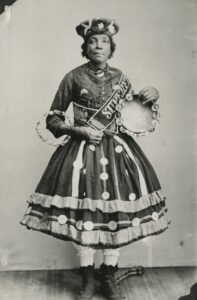 一张黑褐色的照片，照片上是一位中年黑人妇女露西·米切尔，她穿着一件黑色长裙，裙摆上有大大的白色圆纽扣，上面缝着宽阔的白色条纹。她还有一顶有趣的贝雷帽样式的帽子，一条刺绣腰带，一个花环和一个手鼓。