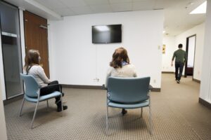 在一间办公室里，为了保持社交距离，两个人坐在椅子上，而一名临床医生在他们前面的走廊上走着。