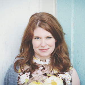 Norma MacDonald, a young white woman with longer reddish hair, wearing a dress with a bold floral pattern.