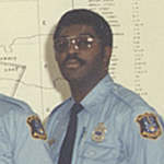 一张Brian Johnston的旧照片，一名穿着黑色镶边眼镜，穿着蓝色衬衫，海军领带和徽章的警察制服。