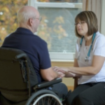 一名白人护士坐在椅子上与轮椅上的一名白人老人谈话，在长期护理设施里面。