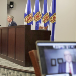 在新冠肺炎疫情通报会上，远处的休斯敦总理坐在桌前，电脑屏幕上的前景是罗伯特·斯特朗博士