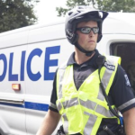 一名白人警察，戴着墨镜，戴着头盔，穿着醒目的背心站在警车前