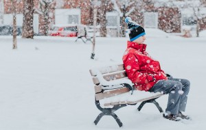 冬天，一个穿着红色夹克、戴着黑色帽的年轻人悲伤地坐在公园的长椅上。下雪了，雪覆盖了地面。他周围的一切几乎都是白色的。背景中隐约可见一条模糊的街道