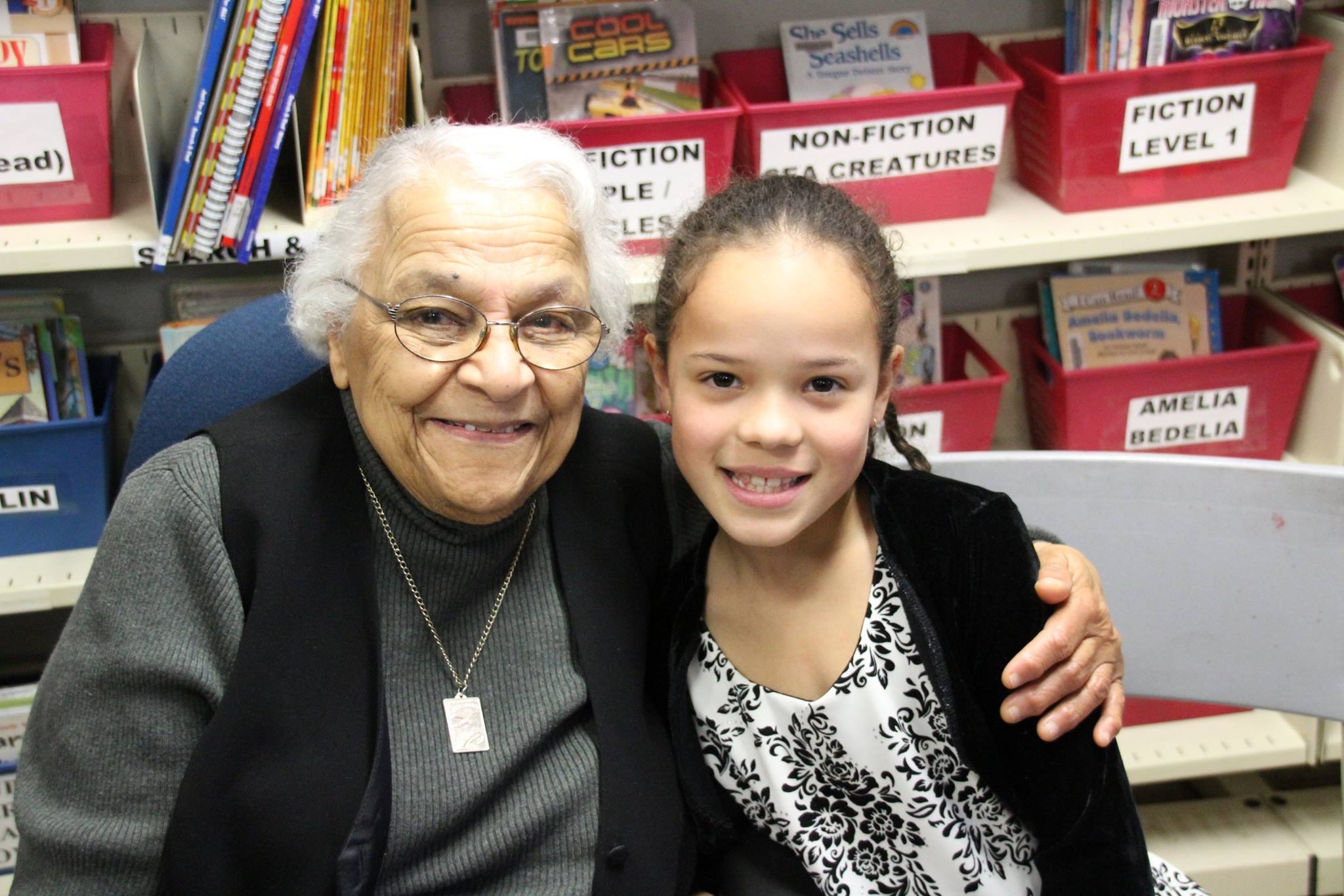 在学校图书馆，当他们对着镜头微笑时，年长的黑人女士用左臂环抱着一个黑人小女孩