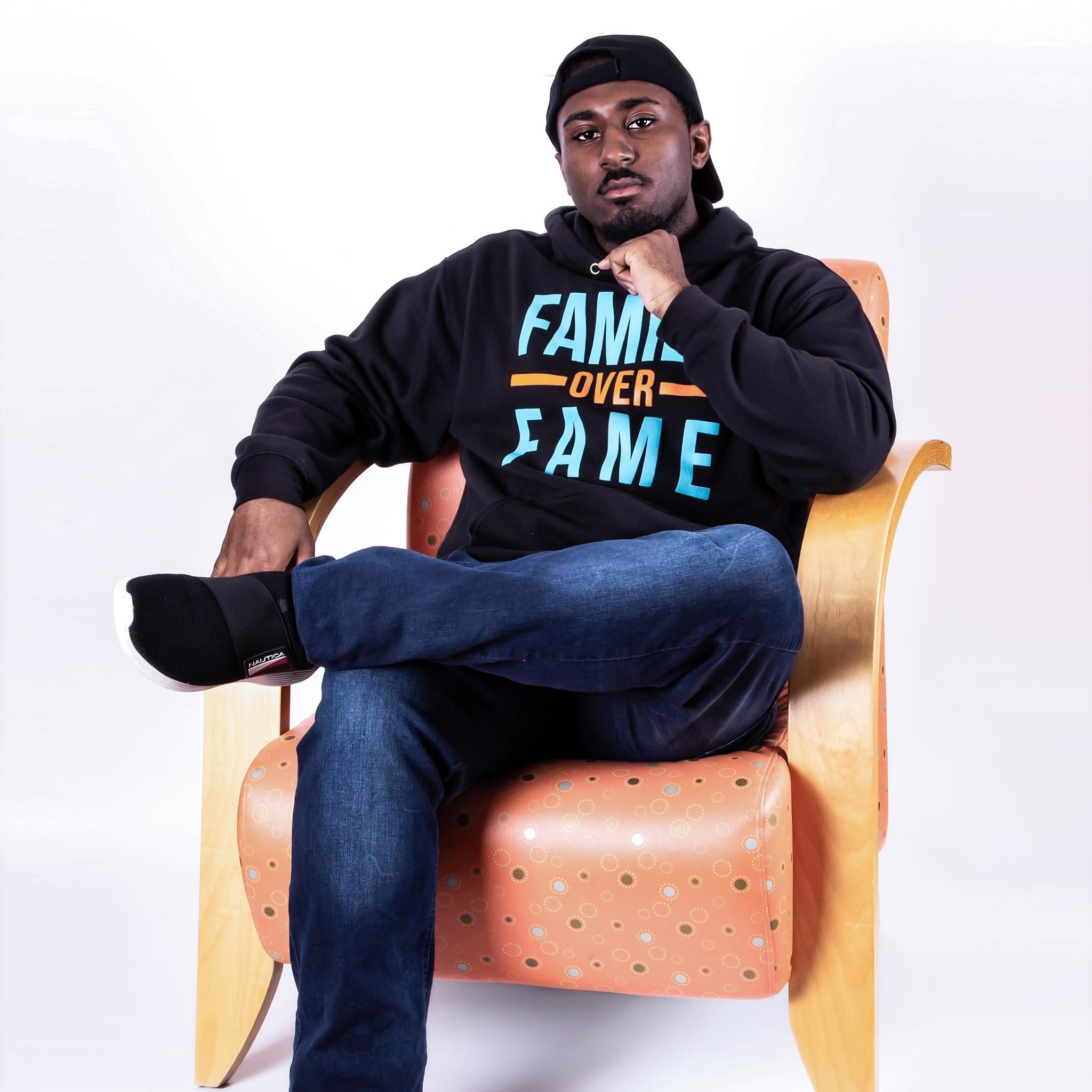 一名身穿蓝色牛仔裤、黑色“Family Over Fame”卫衣的黑人男子双腿交叉坐在扶手椅上