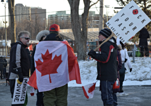Protestors hold signs against public health mandates and chat with a man draped in a Canadian flag.