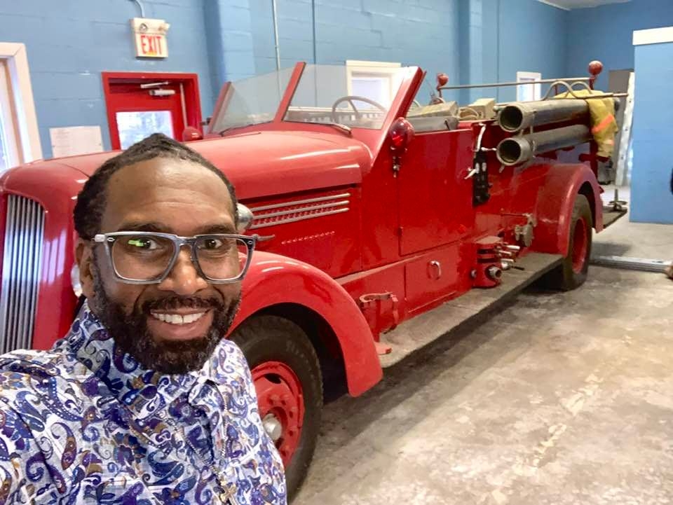戴眼镜的黑人微笑着，背景是一辆老式红色消防车