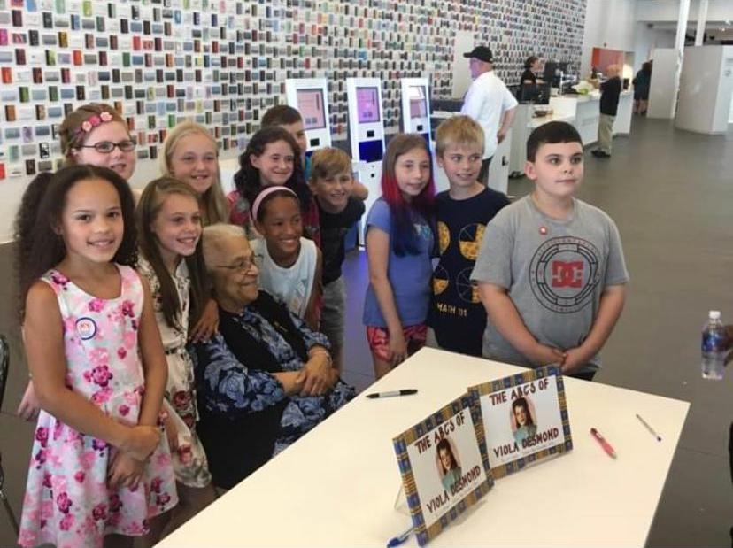 一群孩子和一位年长的黑人女士在图书馆的两本儿童读物后面合影。