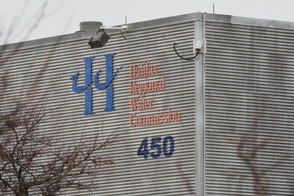 A building will corrugated steel siding is seen on a snowy, grey day. On the side of the building is a blue H with a wave through it, Halifax Water's logo. Next to the H, written in orange, 