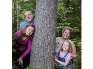 摆在树林里的一个家庭。在右侧，一个微笑的妈妈，短发有她的手在年轻的女儿的肩膀上，一个微笑的年轻白肤金发的女孩戴着整体和一件粉红色的衬衫。到树干的左边，一件紫色衬衫的一个微笑的老女孩倾斜到树上，而她的父亲在她的笑容后面。他戴着眼镜和一件蓝色和白色的衬衫。