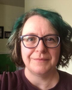 A white woman with glasses and short bobbed style hair with green highlights.