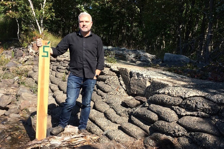 一名穿着黑色衬衫和牛仔裤的男子站在一堵石墙前，手放在一块木板上。板子顶部有绿色胶带，表示5'。这是一个阳光灿烂的日子。