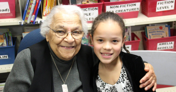 在学校图书馆，当他们对着镜头微笑时，年长的黑人女士用左臂环抱着一个黑人小女孩