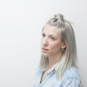 Gabrielle Papillon是一个有着棕色大眼睛的金发女人。她穿着一件浅蓝色的衬衫，靠着一堵浅灰色的墙站着。