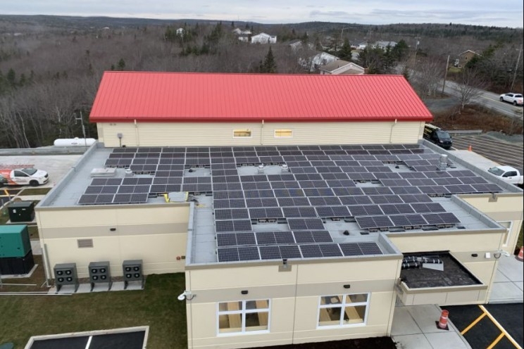 Solar panels are seen on part of the roof of a beige building on a grey day. Another roof in the back of the building is red.
