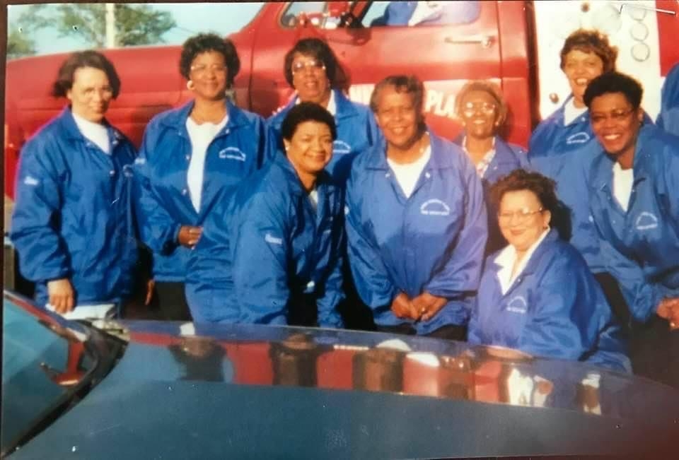 9名身穿蓝色夹克的黑人妇女微笑着，红色消防车是背景。