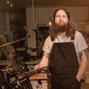 Alex MacAskill，一个留着长发和胡子的年轻白人，站在他的印刷机旁