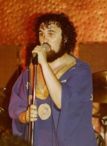 A man with a beard wearing a blue tunic and singing.