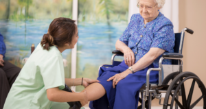 一名身穿薄荷绿色手术服的妇女微笑着望着坐在轮椅上扶着她腿的老年女性。