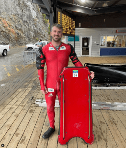 一个穿着红色连体衣的男人微笑着站在一个木制平台上，手里拿着一个红色的骷髅雪橇。那个男人是中年白人