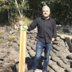 A man stands beside a stone wall, with his hand on a wooden marker with the number 5 on it.
