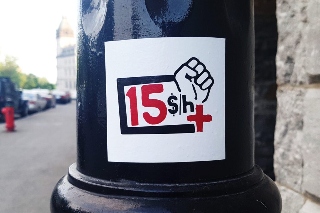 A sticker on a black pole is seen on the side of a street. In the sticker is '15' in red, with '$/h' in black, and then '+' in red, along with a raised fist outlined in black.