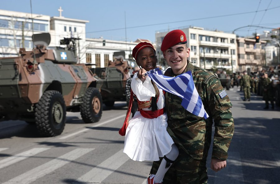 一个身穿军装的白人男子抱着一个身穿传统希腊服装的黑人儿童。都是面带微笑。在背景中可以看到军车，这是阅兵的一部分。