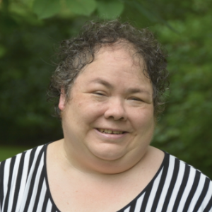 A smiling woman with short curly dark hair wearing a black and white striped top, in front of dark green leafy trees