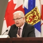 一张戴着眼镜的白人男子坐在桌子前的照片，身后是新斯科舍省和加拿大国旗。