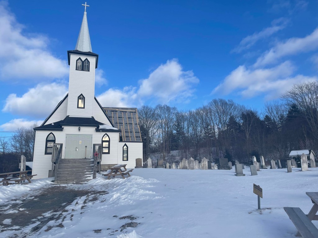 一座带尖塔的白色教堂坐落在蓝天下的小山上。同一处地产上还有一座墓地。