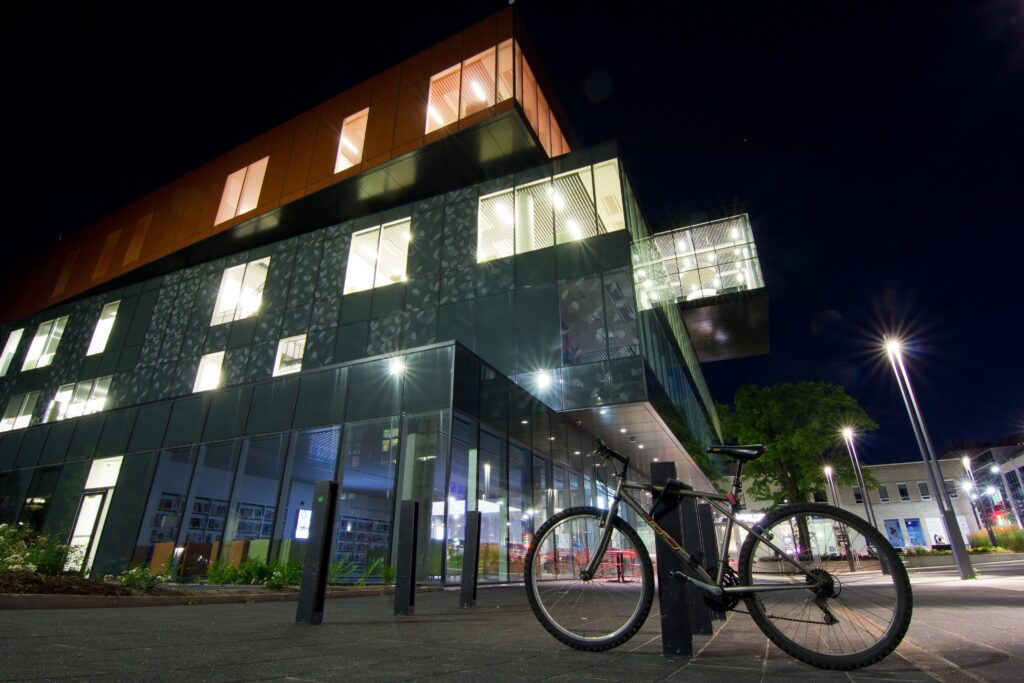 a large glass building is seen from a low angle at night. There's a bicycle locked up in the foreground, and the lights in the building's upper floors are on.