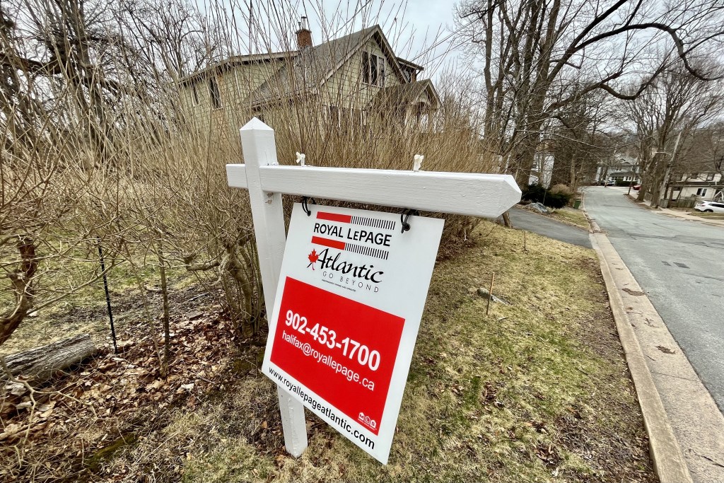 On a grey day, a red and white real estate sign stands out against a brown and green early spring scene. The sign says 
