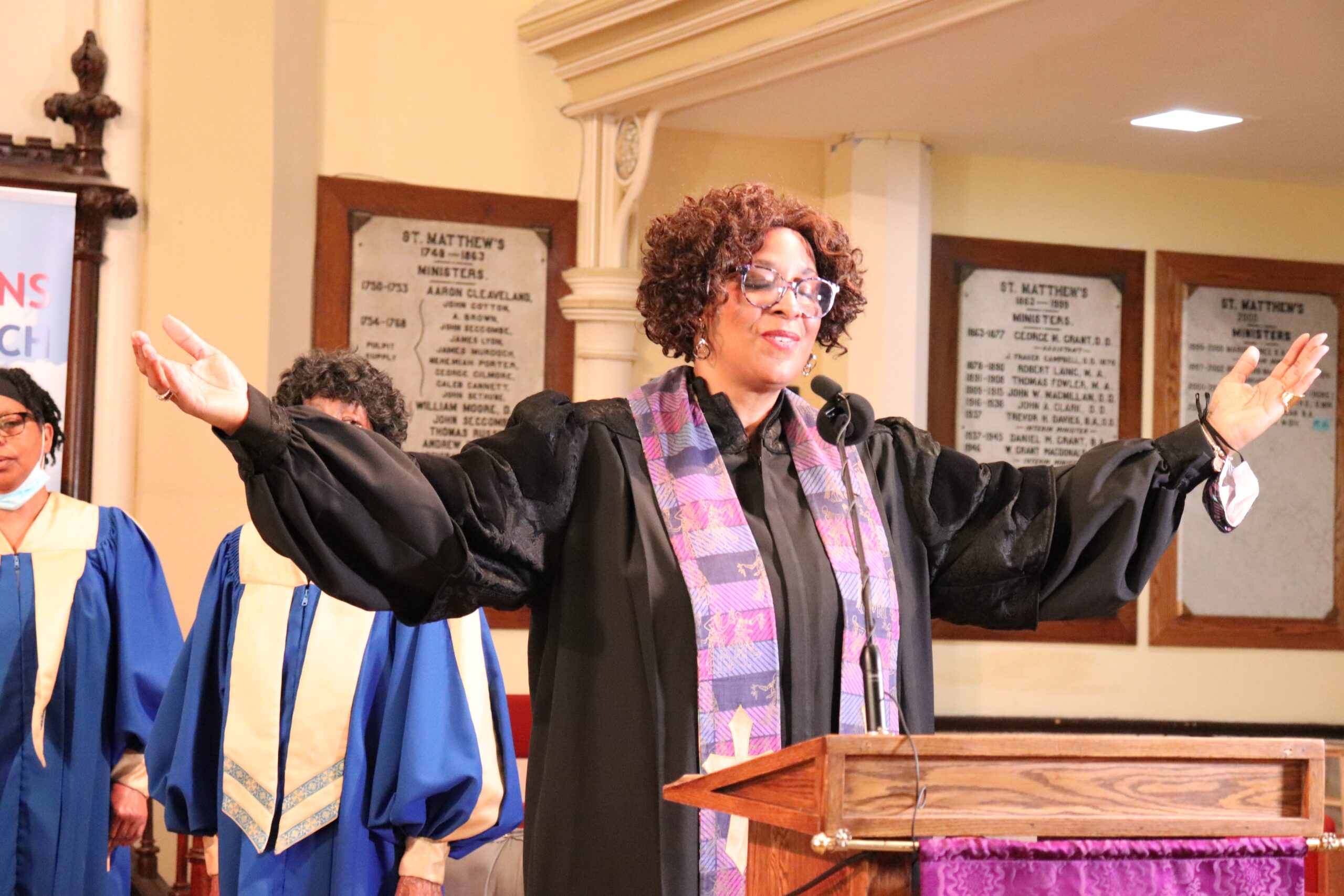 黑人女牧师戴着眼镜，穿着黑色长袍，闭上眼睛，微笑着，抬起手臂和双手放在身体两侧