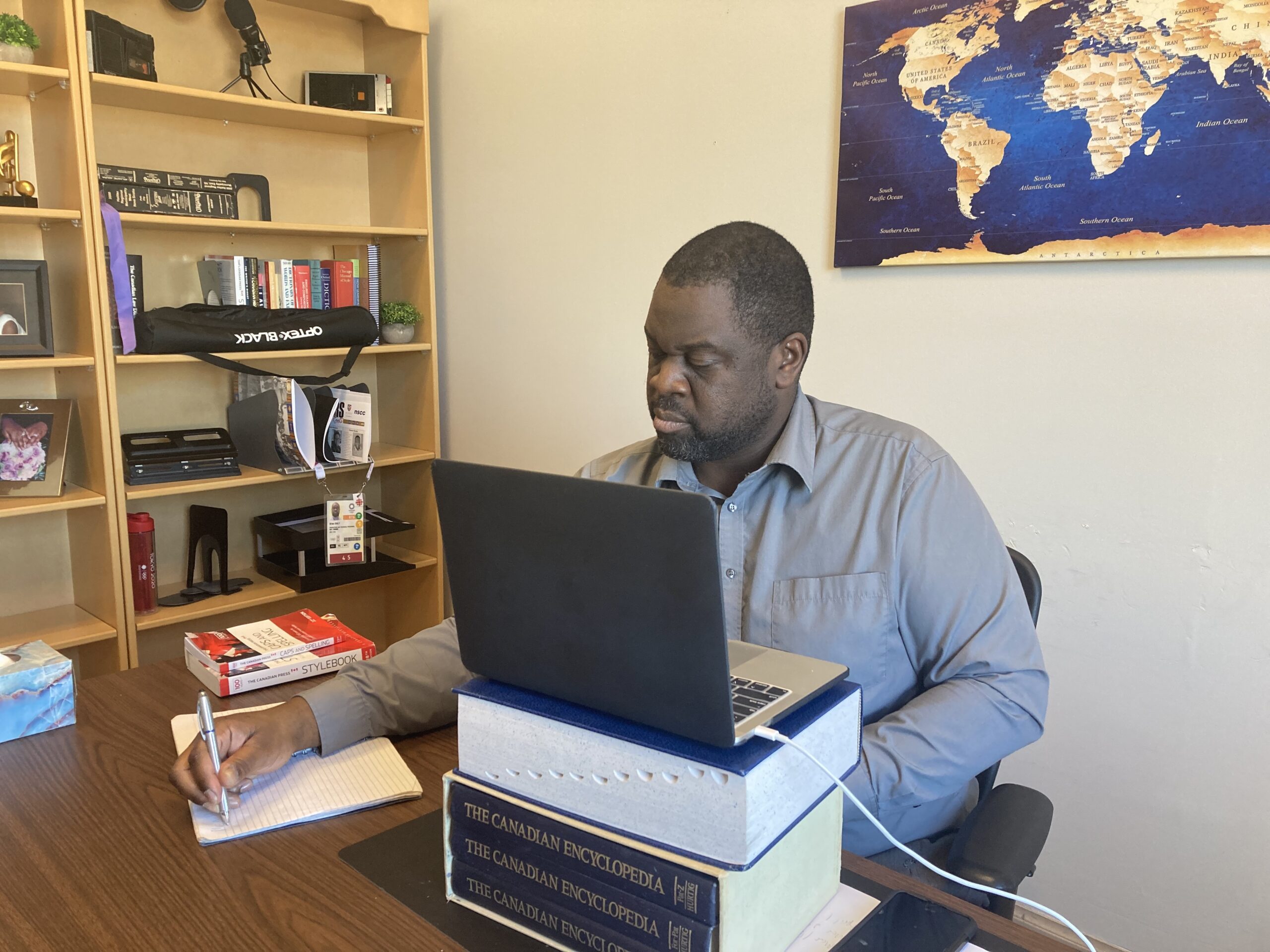 一个穿着灰领衬衫的黑人坐在桌前，打开笔记本电脑，在一摞书上写着便条