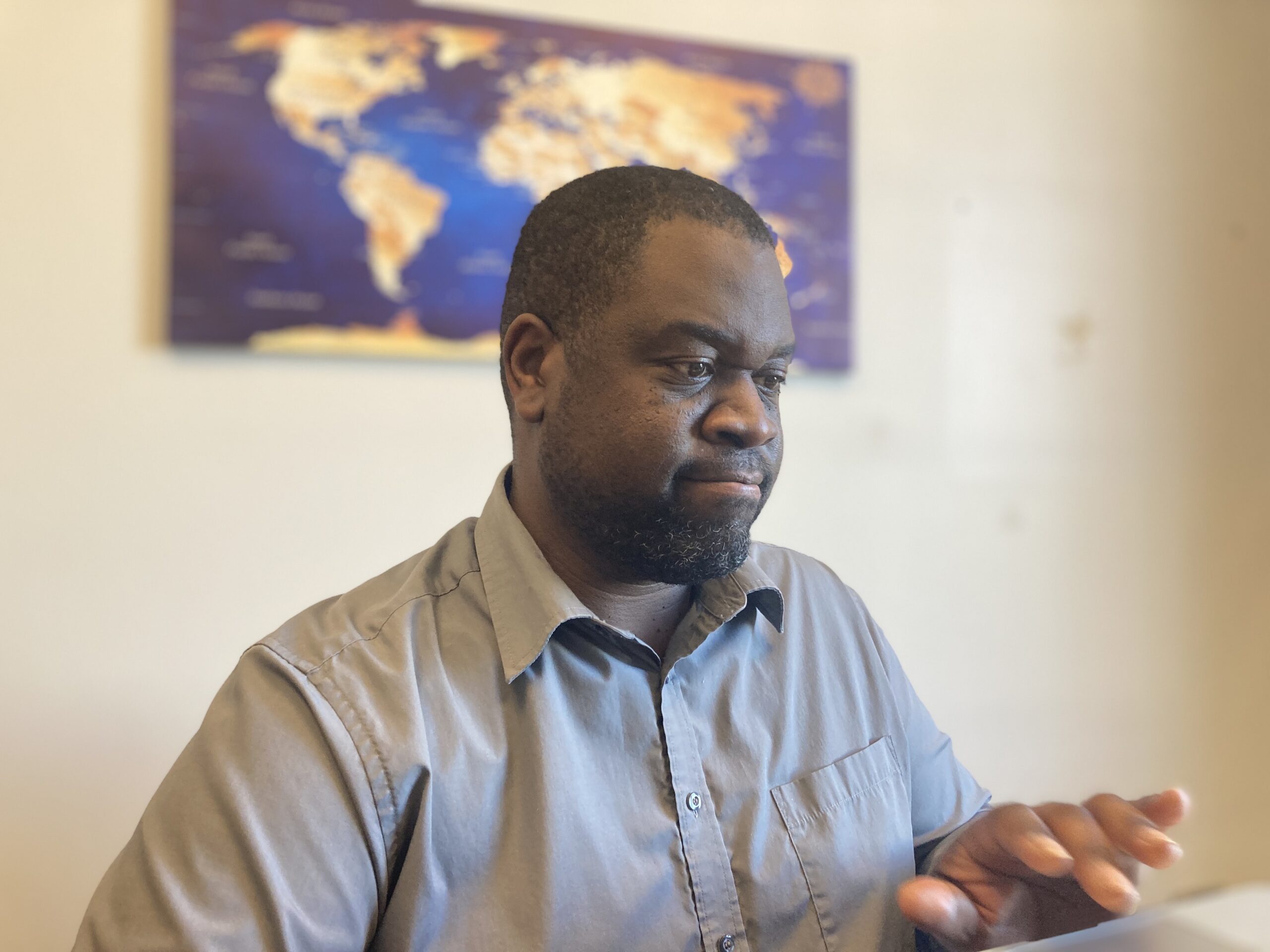 穿着灰领衬衫的黑人男子在笔记本电脑前打字，背景是墙上模糊的世界地图