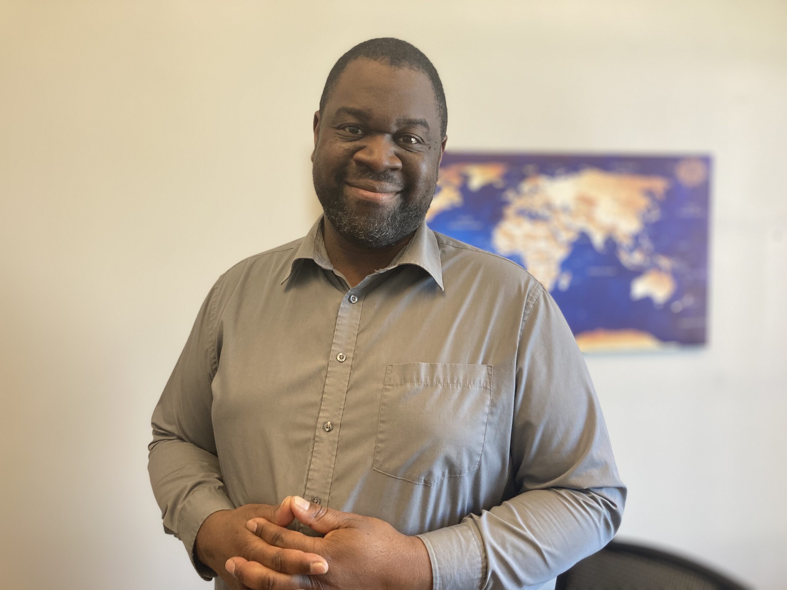 身穿灰领衬衫的黑人男子对着镜头微笑，墙上的背景是模糊的世界地图