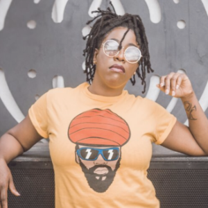 A young Black woman wearing sunglasses and a pale orange t-shirt with a cartoon of a Black man's face on it