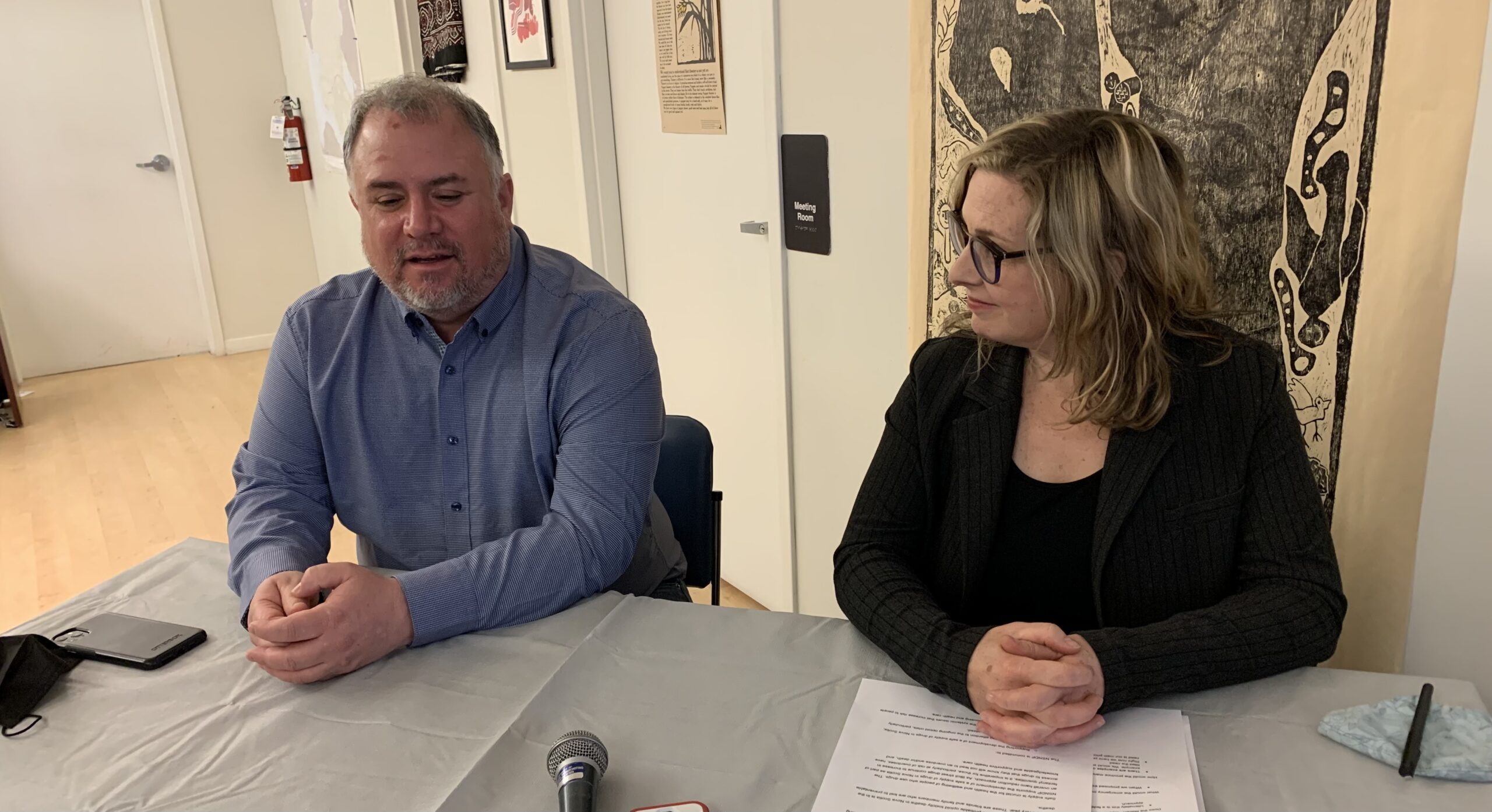 A man with a grey beard and hair wearing a pale blue shirt and a woman with shoulder length light brown hair, dark rimmed glasses, and a black suit sit at a desk.