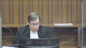 A judge with glasses and a black robe behind a plexiglass screen in a courtroom